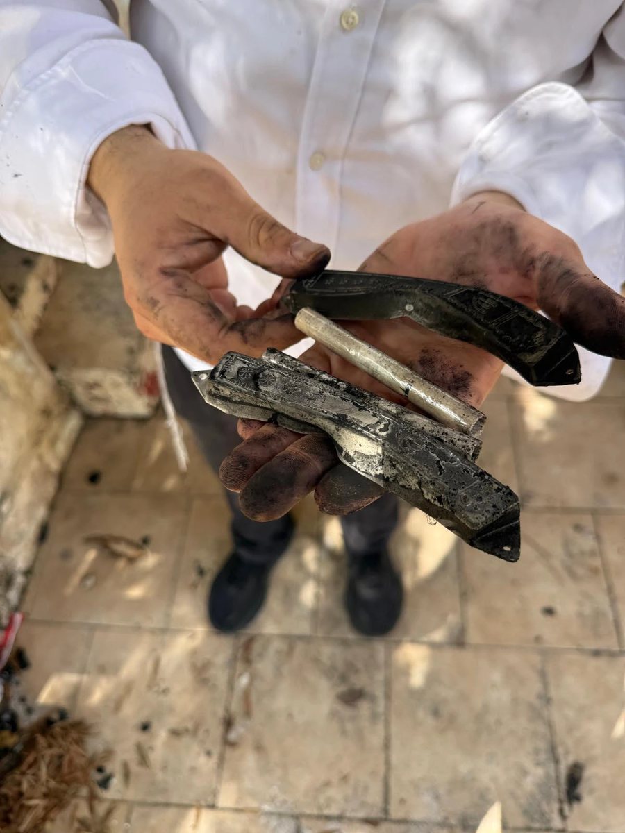 Soot-covered hands holding mezuzah cases blackened and melted by the fire, beside a clean, intact scroll tube