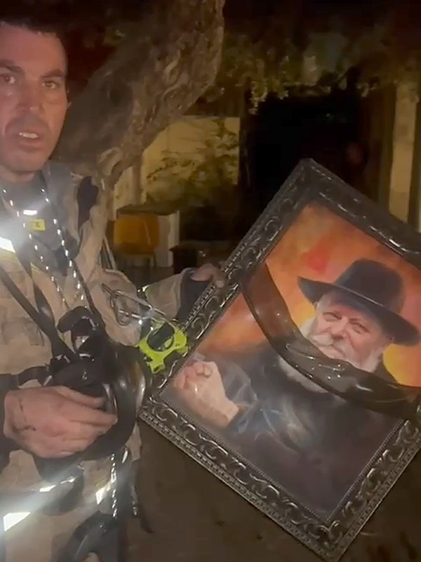 A firefighter in turnout gear holds up the framed portrait of the Rebbe at night, after the fire